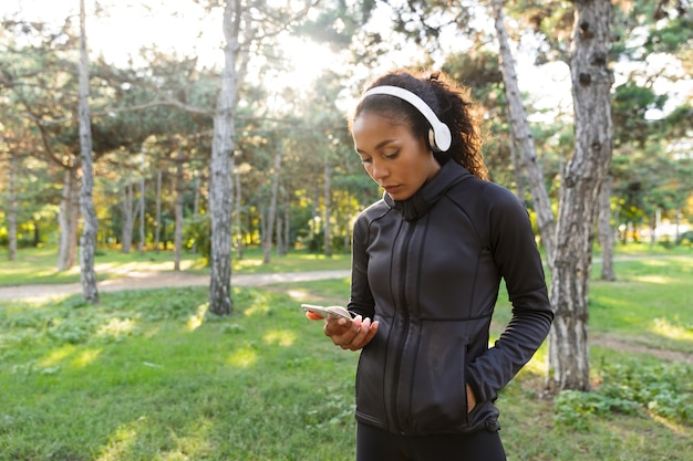 Imagen de una mujer bonita de 20 años vistiendo un chándal negro y auriculares, con teléfono móvil mientras camina por el parque verde