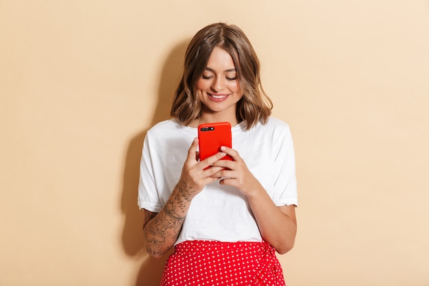 Imagen de mujer bonita en falda roja sonriendo y sosteniendo smartphone aislado sobre pared beige