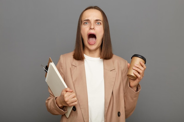 Imagen de una mujer caucásica de cabello castaño conmocionada y desesperada con una chaqueta que sostiene un portapapeles con documentos y un café para llevar posando aislado sobre un fondo gris