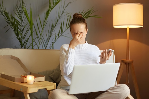 Imagen de una mujer caucásica exhausta e insalubre con suéter blanco sentada en un sofá y trabajando en una laptop sosteniendo un teléfono inteligente en las manos frotándose los ojos