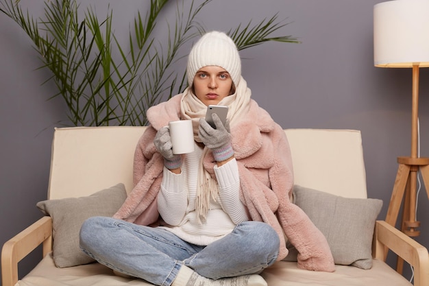 Imagen de una mujer caucásica triste y disgustada con ropa abrigada y envuelta en una bufanda sentada en un sofá en la habitación con una taza de té caliente sosteniendo un teléfono móvil mirando la cámara