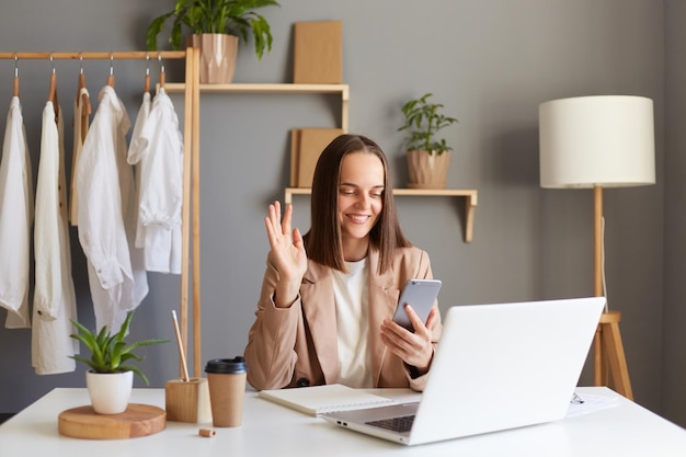 Foto imagen de una mujer con chaqueta trabajando en una laptop en la oficina en casa contra la ropa colgada en un estante con un descanso haciendo una videollamada con un amigo de un socio comercial agitando la mano mostrando un gesto de saludo