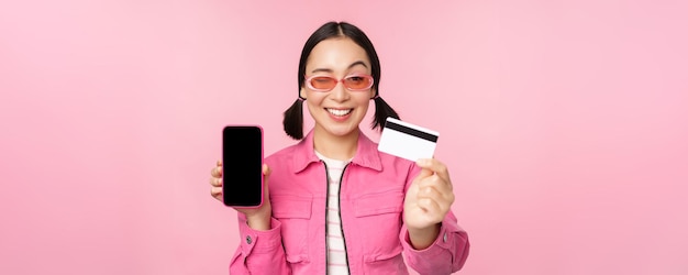 Imagen de una mujer coreana sonriente que muestra la tarjeta de crédito y la interfaz de la aplicación del teléfono inteligente de la pantalla del teléfono móvil pagando compras en línea sin contacto de pie sobre fondo rosa