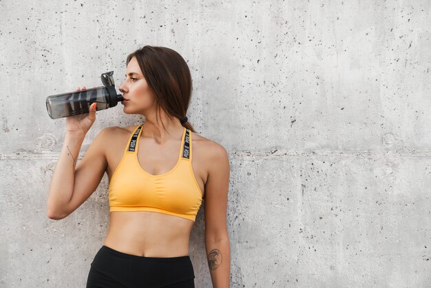 Imagen de mujer delgada en ropa deportiva bebiendo agua de una botella de plástico mientras está parado sobre un muro de hormigón al aire libre