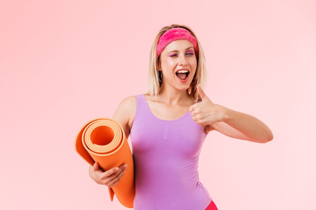 Imagen de mujer deportiva encantada en diadema sosteniendo mat y gesticulando pulgar hacia arriba aislado sobre pared rosa