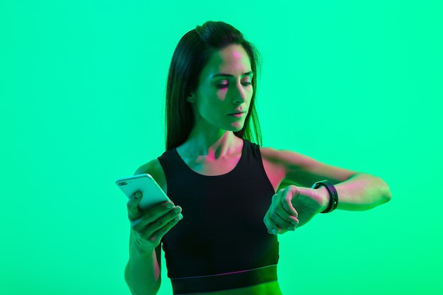 Imagen de una mujer deportiva fuerte bastante concentrada joven aislada sobre pared azul con luces de neón brillantes con teléfono móvil mirando el reloj.