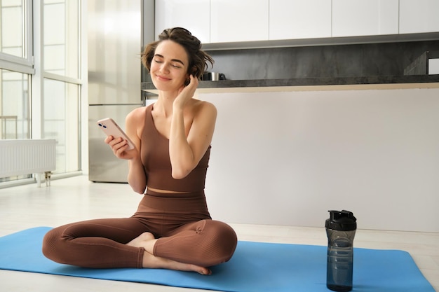 La imagen de una mujer deportiva en ropa deportiva se sienta en una alfombra de yoga en casa sostiene que el teléfono inteligente usa la búsqueda de la aplicación de entrenamiento