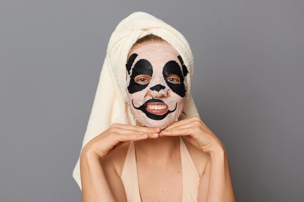 Imagen de una mujer divertida con la parte superior y una toalla en la cabeza aislada sobre fondo gris parada con una máscara de panda en la cara haciendo procedimientos de cuidado de la piel de cosmetología mantiene las manos debajo de la barbilla