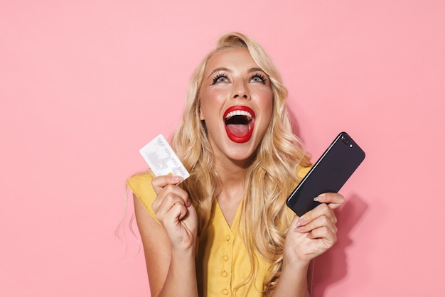 Imagen de mujer emocionada con largo cabello rubio sonriendo mientras sostiene el teléfono celular y la tarjeta de crédito aislada sobre la pared rosa