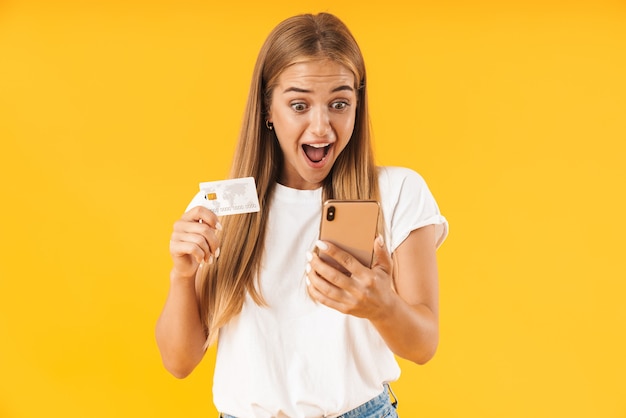 Imagen de mujer emocionada en ropa básica sonriendo mientras sostiene el teléfono inteligente y la tarjeta de crédito aislada sobre la pared amarilla