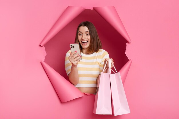 Imagen de una mujer emocionada y sorprendida con un pantalones a rayas posando en un avance de fondo rosa de pie con bolsas de compras y un teléfono inteligente revisando el reembolso