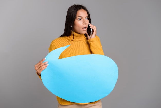 Foto imagen de una mujer emocionada sorprendida posando aislada sobre la pared de la pared gris sosteniendo bocadillo hablando por teléfono.