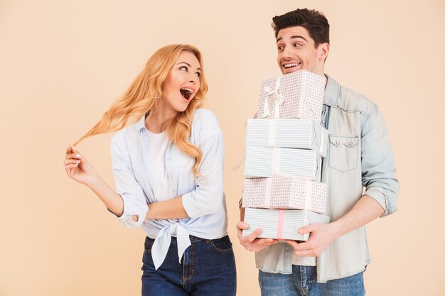 Imagen de una mujer encantadora regocijándose y expresando sorpresa mientras un hombre guapo sostiene un montón de cajas de regalo, aisladas sobre una pared beige