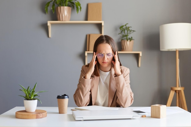 Imagen de una mujer enferma cansada y sobrecargada de trabajo con el pelo castaño y una chaqueta beige trabajando en la oficina trabajando duro sintiendo fatiga y terrible dolor de cabeza frunciendo el ceño masajeando las sienes