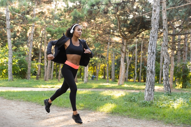 Imagen de mujer estadounidense de 20 años vistiendo chándal negro y auriculares haciendo ejercicio, mientras corre por el parque verde