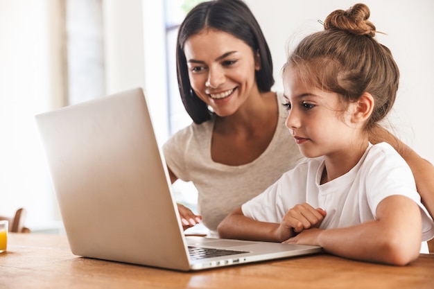 Foto imagen de mujer de familia atractiva y su pequeña hija sonriendo y usando la computadora portátil juntos mientras está sentado a la mesa en el apartamento