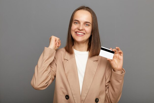 Imagen de una mujer feliz y atractiva con el pelo castaño y una chaqueta beige posando aislada sobre un fondo gris ganando la lotería mostrando su tarjeta bancaria con el puño cerrado celebrando