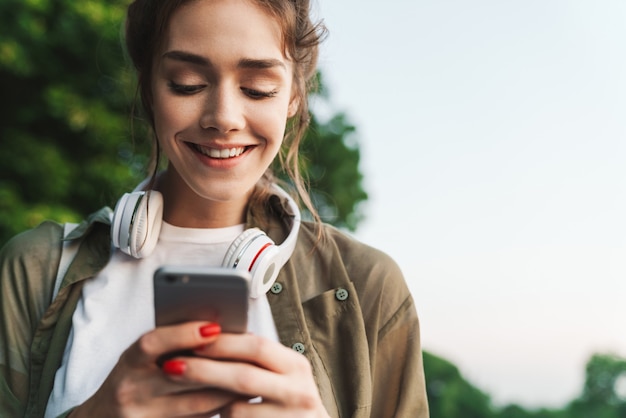 Imagen de mujer feliz con auriculares sobre su cuello sosteniendo smartphone y caminando en el parque verde