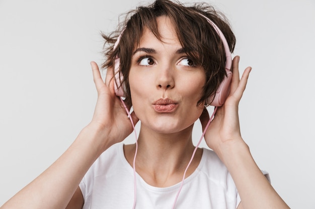 Imagen de una mujer feliz bastante emocionada joven hermosa que presenta aislada sobre la pared blanca que escucha música de la pared con los auriculares.