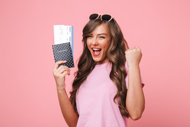 Imagen de mujer feliz con hermosas cerraduras marrones regocijándose de sus vacaciones mientras sostiene pasaporte y boletos de avión, aislado sobre fondo rosa