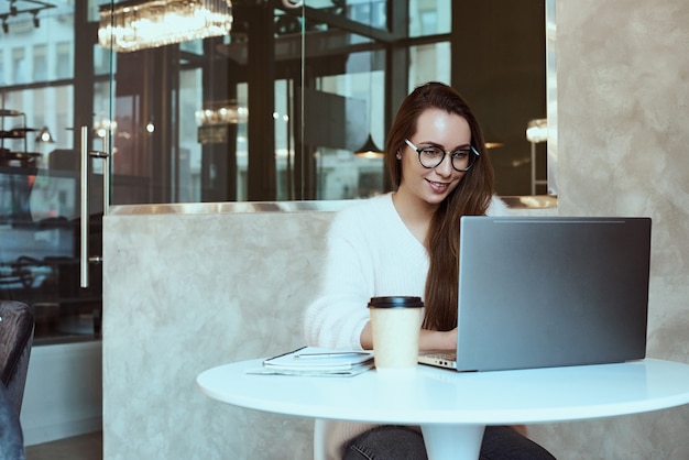 Imagen de la mujer feliz que usa la computadora portátil mientras que se sienta en el café. Merican mujer sentada en una cafetería y trabajando en equipo portátil.