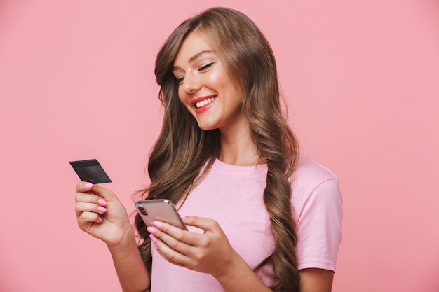 Imagen de mujer feliz en ropa casual sosteniendo teléfono celular y tarjeta de crédito mientras compra en internet, aislado sobre fondo rosa