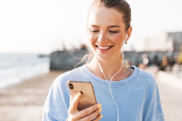 Imagen de una mujer de fitness joven alegre positivo al aire libre en la playa mediante teléfono móvil escuchando música con auriculares.