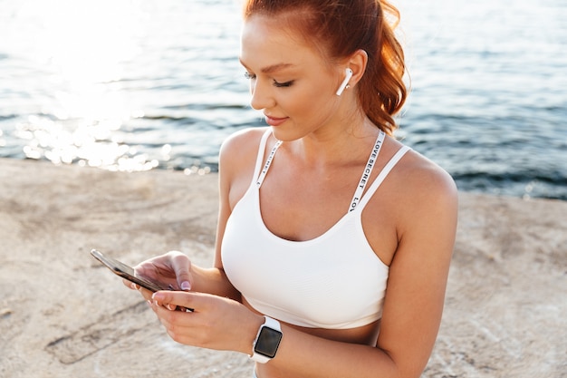 Imagen de mujer fitness pelirroja joven positiva al aire libre en la playa por la mañana escuchando música con auriculares inalámbricos bluetooth mediante teléfono móvil.