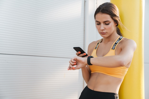 Foto imagen de mujer fitness en ropa deportiva sosteniendo teléfono celular y mirando el reloj de pulsera al aire libre