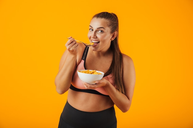 Imagen de mujer gordita deportiva en chándal comiendo copos de maíz con leche para el desayuno, aislado sobre fondo amarillo