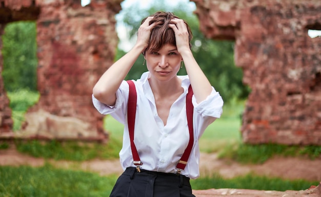 Imagen de una mujer hermosa y elegante con una camisa blanca, pantalones negros y tirantes rojos sentada en un parque con el telón de fondo de un edificio destruido El concepto de estilo y moda