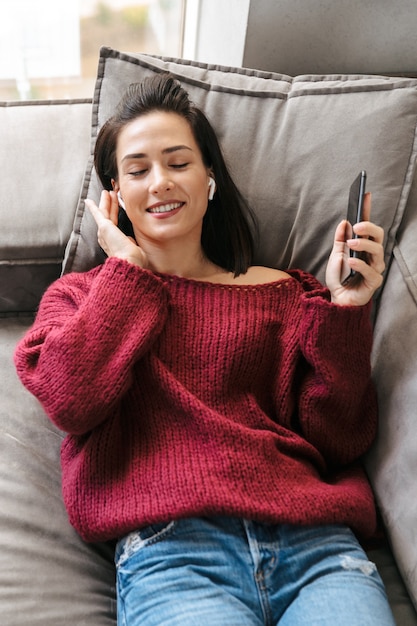 Imagen de una mujer hermosa en el interior de su casa en el sofá escuchando música con auriculares.