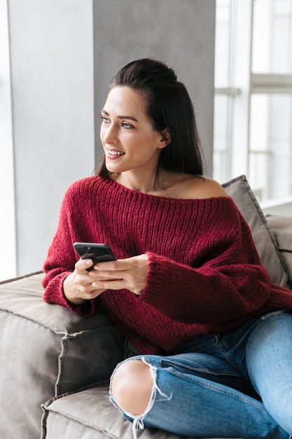 Imagen de una mujer hermosa en el interior de su casa en el sofá mediante teléfono móvil.