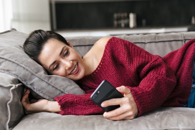 Imagen de una mujer hermosa en el interior de su casa en el sofá mediante teléfono móvil.