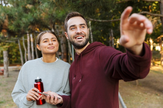 Imagen de una mujer y un hombre felices deportes positivos mediante teléfono móvil al aire libre en el parque natural verde apuntando a un lado sosteniendo una botella con agua.