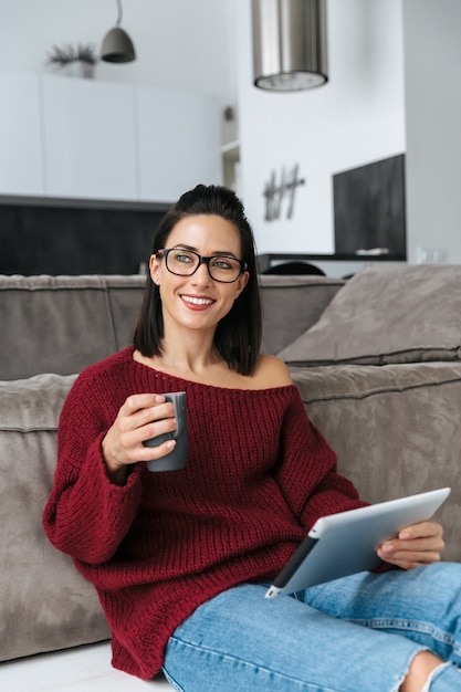 Imagen de una mujer increíble en el interior de la casa en el sofá con tablet PC tomando café.