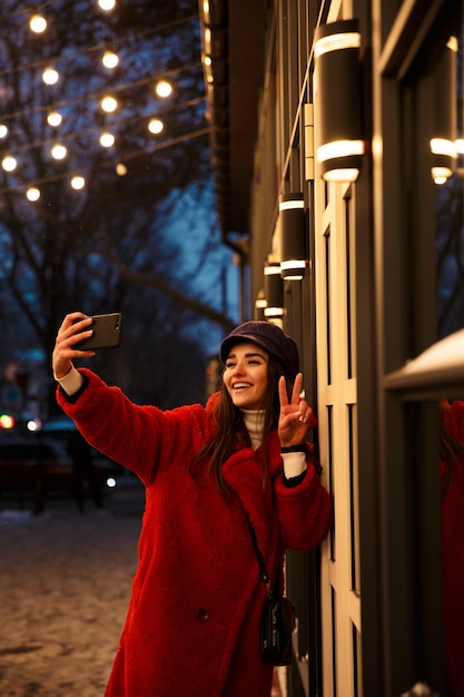 Imagen de una mujer joven y bonita al aire libre caminando en la calle de noche de invierno de nieve tomar selfie por teléfono móvil.