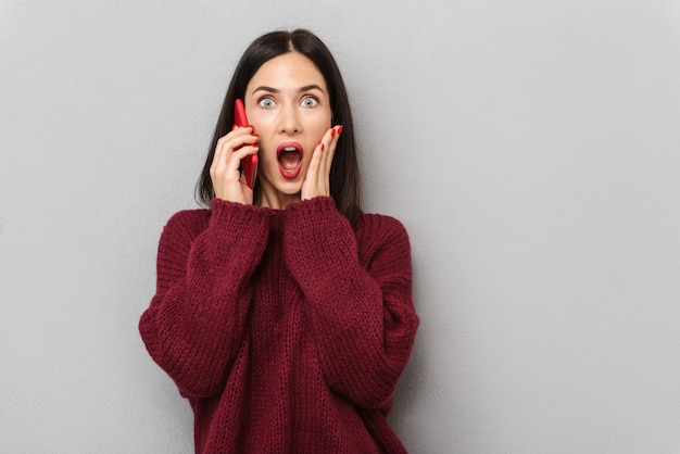 Imagen de una mujer joven y bonita conmocionada vestida con un suéter burdeos hablando por teléfono móvil aislado.