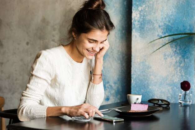 Imagen de una mujer joven y bonita sentada en la cafetería tomando café en el interior con teléfono móvil escuchando música con auriculares.