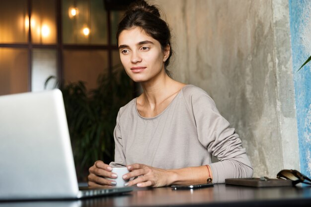 Imagen de una mujer joven y bonita sentada en la cafetería tomando café en el interior usando una computadora portátil.