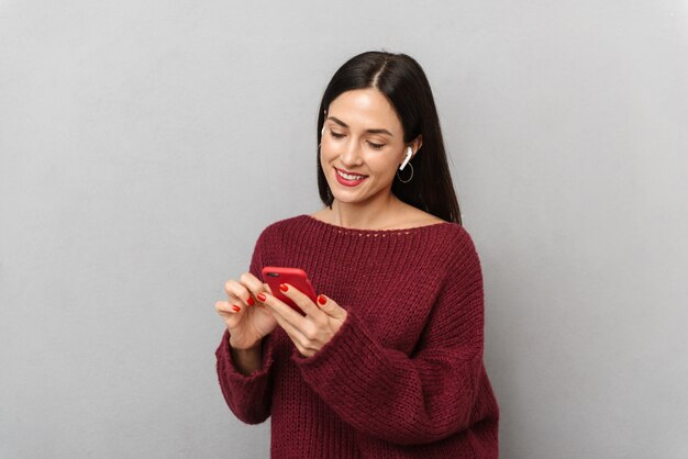 Imagen de una mujer joven y bonita vestida con un suéter burdeos con teléfono móvil aislado escuchando música con auriculares