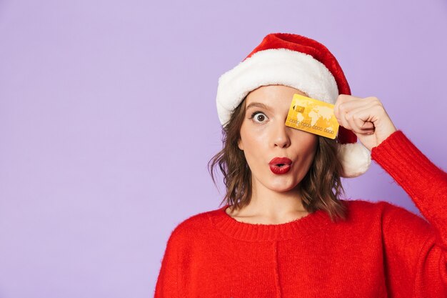 Imagen de una mujer joven conmocionada emocionada con sombrero de navidad aislado sobre pared púrpura con tarjeta de débito.