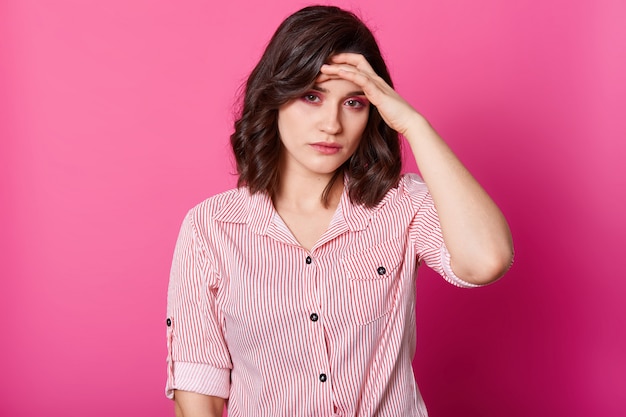 Imagen de mujer joven con dolor de cabeza terrible, mantiene la mano en la frente, necesita tomar medicamentos, foto de estudio de una niña morena en blusa a rayas, aislada sobre rosa