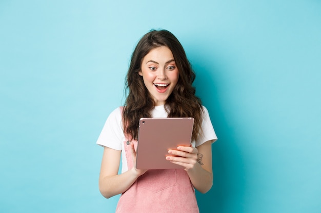 Imagen de una mujer joven emocionada que mira sorprendida a la pantalla de la tableta digital, encontró algo genial en línea, de pie contra el fondo azul