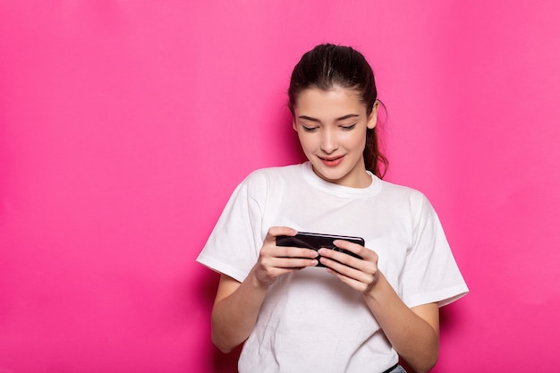 Imagen de mujer joven feliz en camiseta blanca, jugando juegos de teléfono móvil y ganando, regocijándose mientras está parado sobre fondo rosa