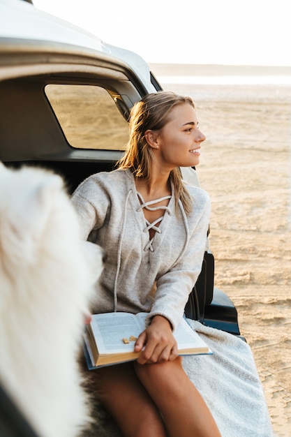 Imagen de mujer joven feliz con perro samoyedo al aire libre en la playa en coche con libro.