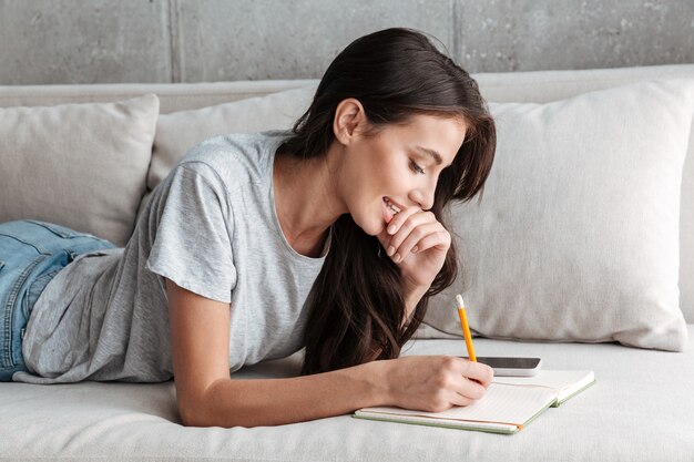 Imagen de mujer joven morena complacida sonriendo y escribiendo notas en el libro diario en el sofá en casa