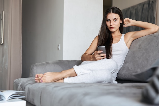 Imagen de una mujer joven muy hermosa sentada en el sofá en el interior de su casa mediante el chat de teléfono móvil.