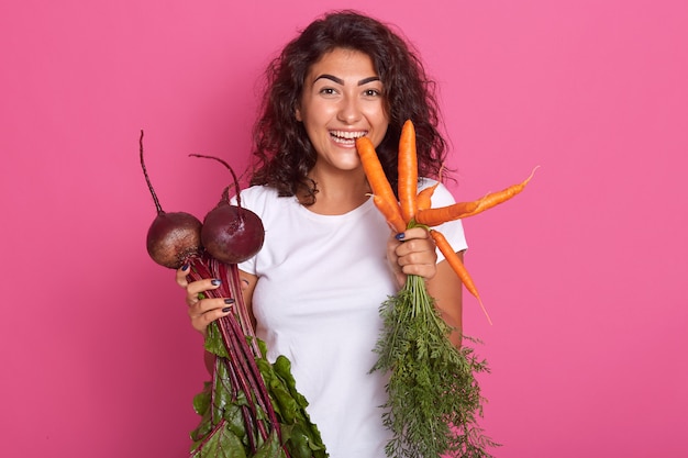 La imagen de la mujer joven con el pelo ondulado oscuro vistió la camiseta casual blanca que sostenía remolachas y zanahorias en las manos, mirando directamente a la cámara, mordiendo la zanahoria. Dieta de alimentos crudos y concepto de alimentación saludable.