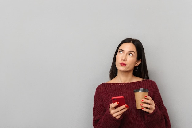 Imagen de una mujer joven pensativa vestida con un suéter de Borgoña usando un teléfono móvil aislado tomando café.